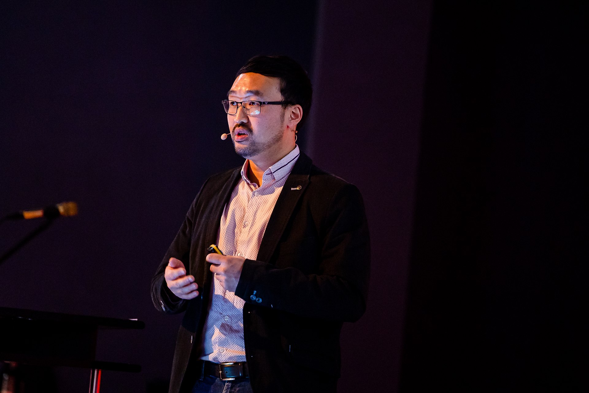 Dr. Seung-Woo Yoo mit Headset-Mikrofon und Fernbedienung auf dunkler Kongressbühne, Rednerpult daneben.